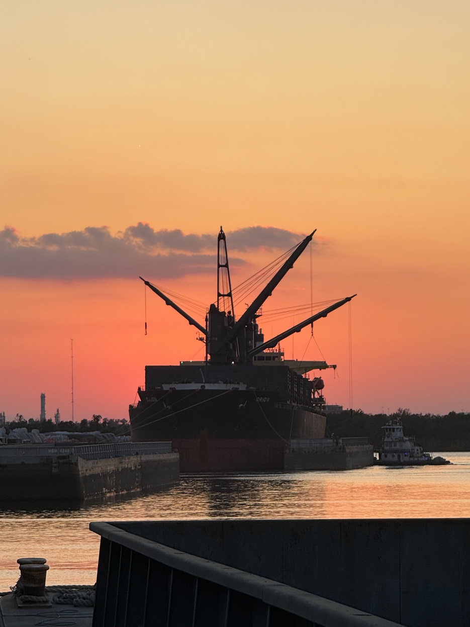 Bulk carrier vessel bow-on silhouette at intense orange sunset, Lake Charles — ship crane rigging lines radiating against glowing sky