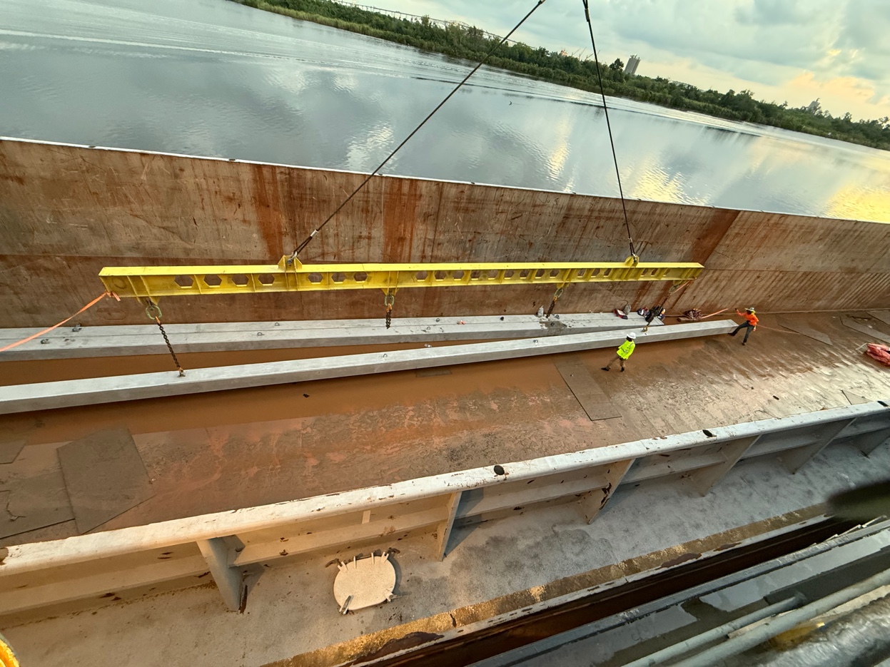 Yellow spreader bar suspended by crane lowering concrete piles, two crew members guiding on deck with river and treeline behind