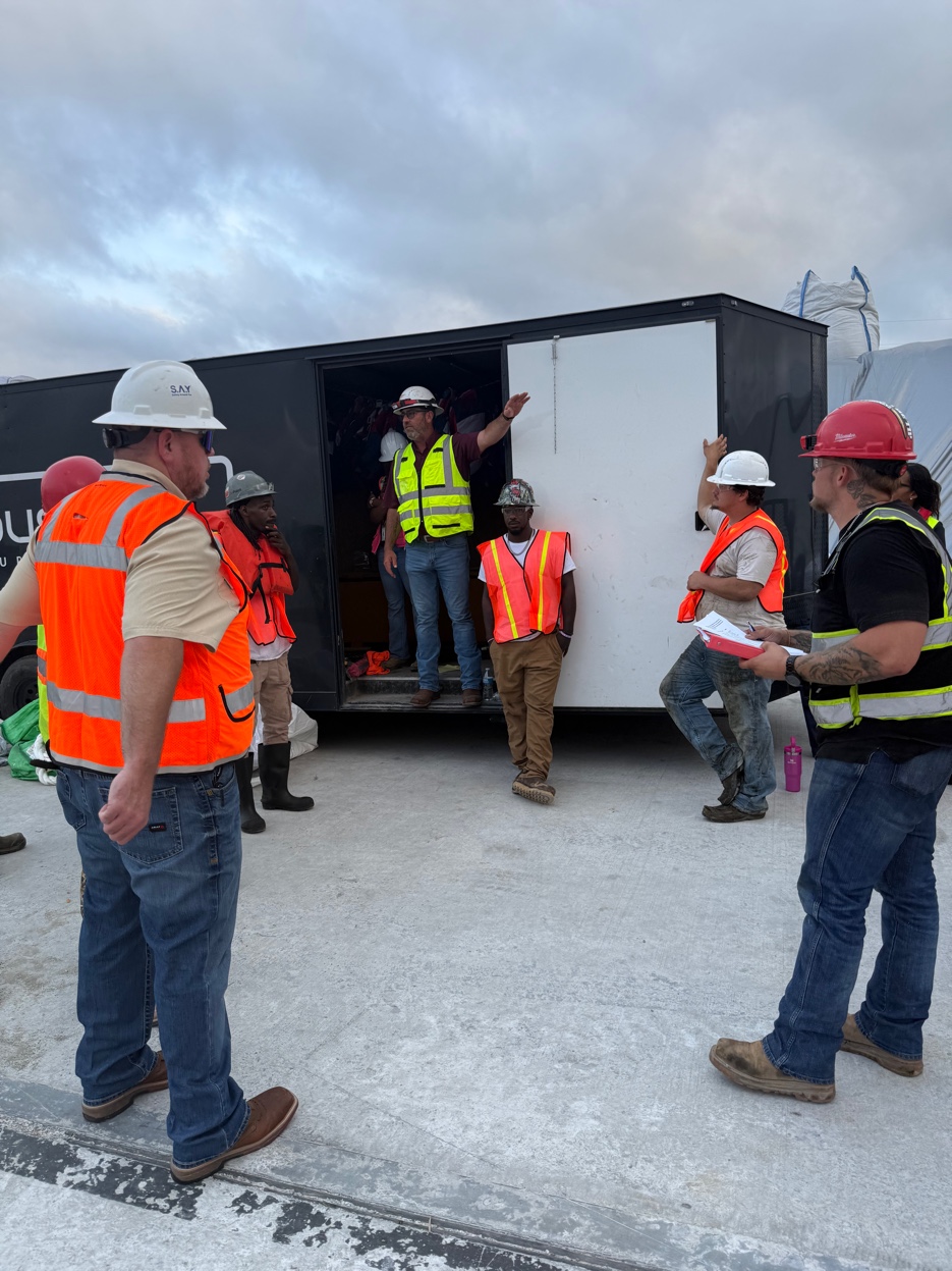 GCS supervisor briefing the crew from the back of a trailer at the job site — crew in hard hats and hi-vis listening to the pre-shift safety talk on deck