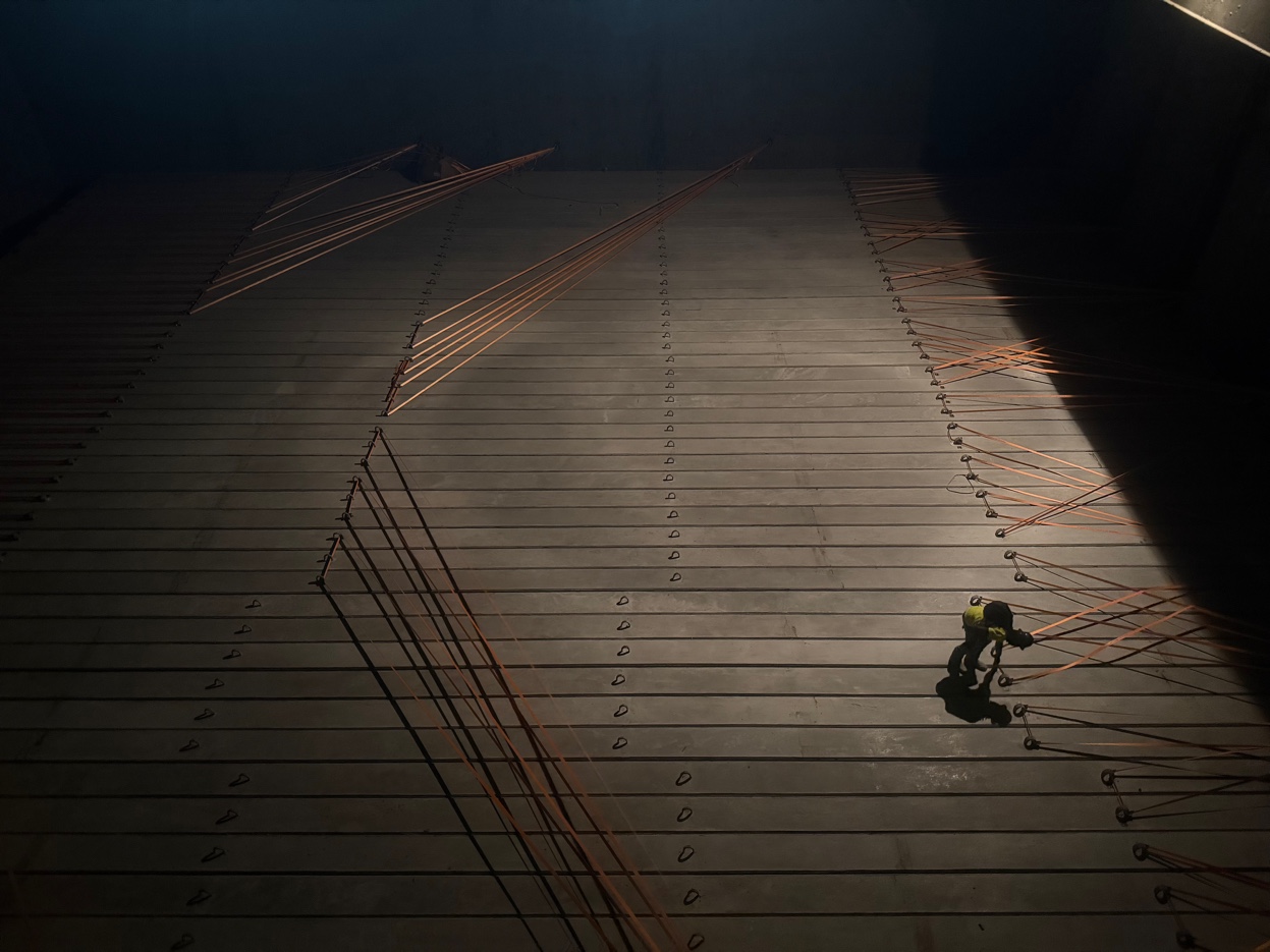 Aerial view of vessel deck at night during GCS discharge operation — lone worker in hi-vis dwarfed by rigging lines fanning across the hold in dramatic light