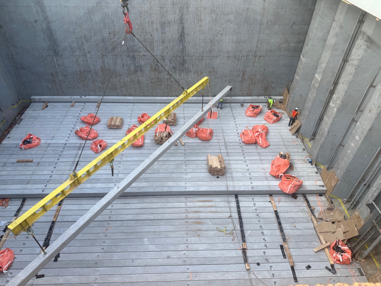 Looking down into vessel hold during MV Atlantic Runner II concrete pile discharge — yellow spreader bar suspended above piles, red rigging bags, crew guides below